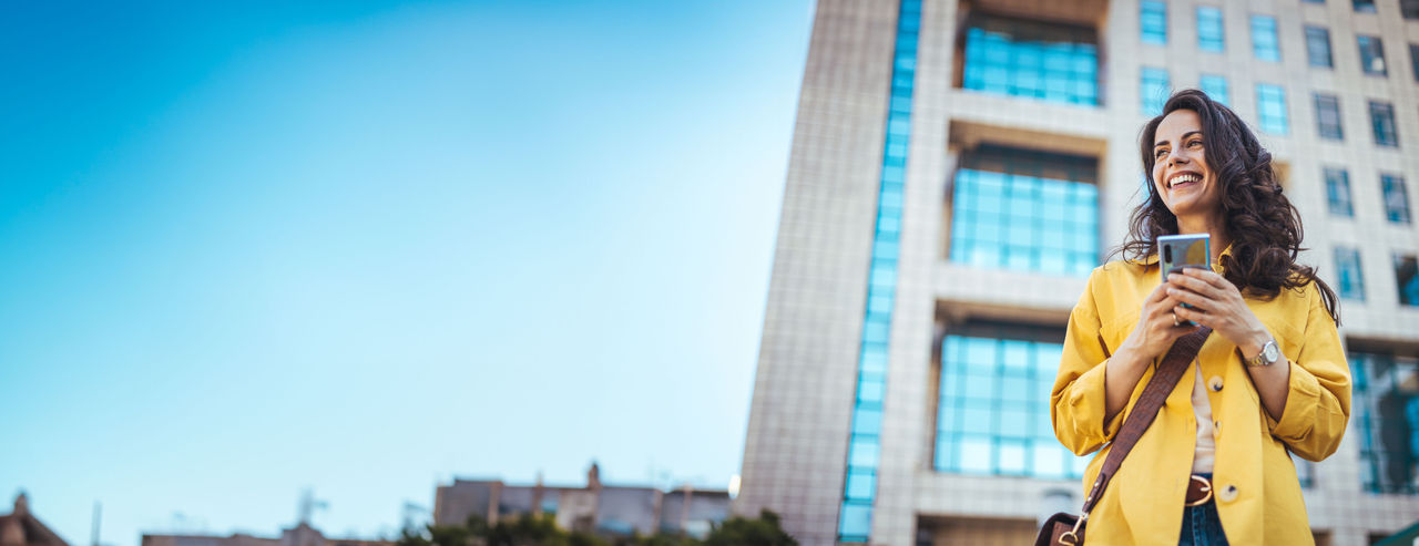 Young adult smiling woman wearing yellow T shirt holding smartphone looking at mobile phone using apps on cellphone technology texting standing on urban city street outside.